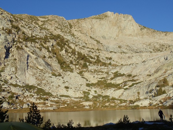 A massive wall of rock, dotted with pine trees, makes up the backdrop for Squaw Lake, in the foreground, as dusk approaches; the sun has set far enough that land before the lake is in shadows, while the lake and slope behind it are yet sunlit