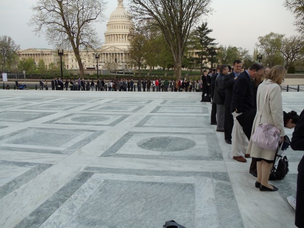 The long line of people extends straight back from the center of the plaza, passes down the steps, then turns left heading south along the sidewalk