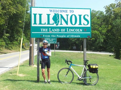 Me standing underneath a "Welcome to Illinois" sign, with my bike leaning against the sign just next to me