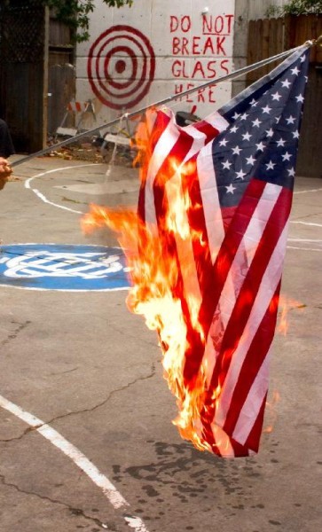 An American flag held, burning, touching the ground